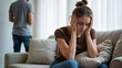 © Juan Mari - A woman sits on a couch, visibly distressed, while a man stands in the background, creating a tense atmosphere. Image is ideal for articles on relationship issues, mental health, and emotional support