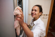 © Taras Grebinets - A smiling woman wearing an apron is cleaning the glass door of a refrigerator with a cloth, embodying household chores, cleanliness, and domestic life in a home kitchen environment.