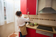 © Taras Grebinets - Young woman in apron reaching for items in a bright kitchen with red cabinets and cooking vegetables on modern stove, representing domestic life, healthy cooking, and culinary skills development.