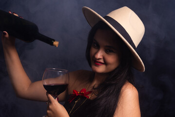 Beautiful woman wearing brown hat and black clothes simulating pouring wine with a bottle and a glass goblet.