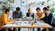 © primopiano - Group of young professionals collaborating at a modern office workspace, reviewing documents and working on laptops, surrounded by plants and office supplies on a wooden table.