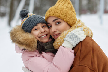 Wall Mural - Portrait of happy young mother and daughter embracing playfully and looking at camera having fun in winter outdoors