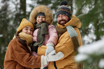 Wall Mural - Waist up portrait of happy young family with little girl in winter forest looking at camera and smiling standing by snowy trees