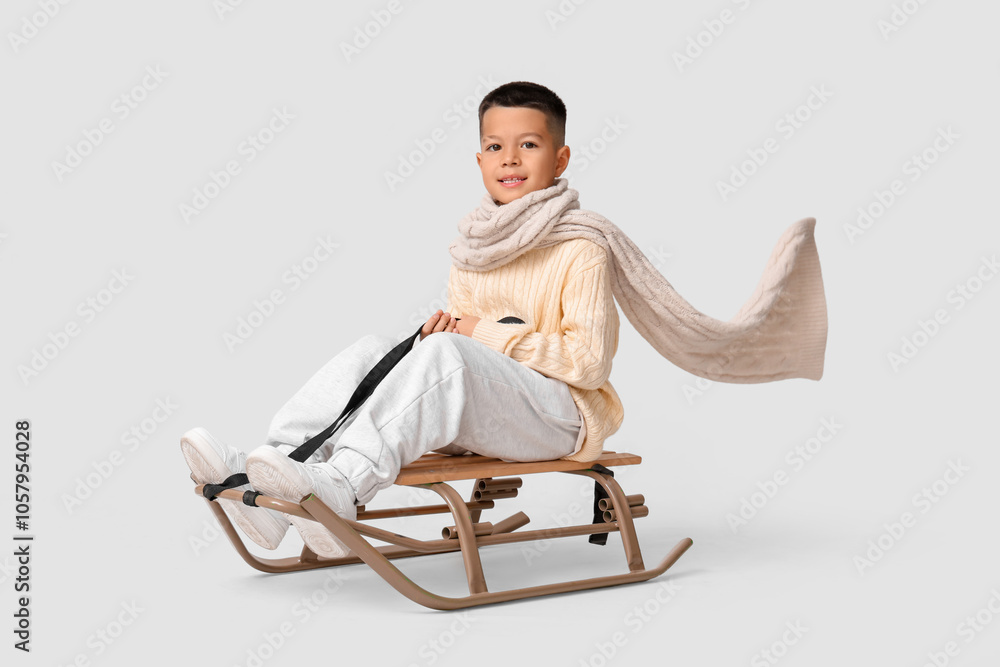 Cute little Asian boy in warm clothes sitting on sledge against grey background
