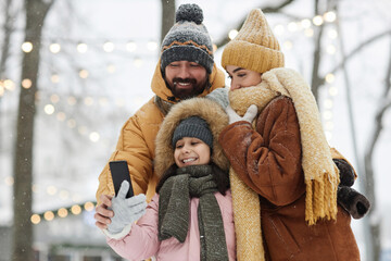 waist up portrait of happy young family with daughter holding phone and taking selfie photo outdoors