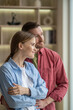 © DimaBerlin - Contented loving man hugging smiling woman standing together at home daydreaming looks at panoramic window. Pleased family couple with spending leisurely time, visualizing future, enjoys calm weekend.