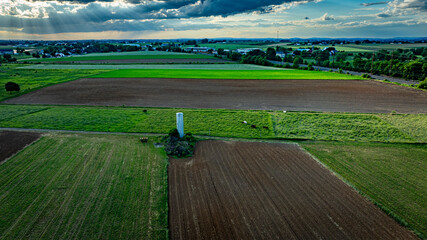 Wall Mural - The lush farmland showcases a patchwork of green and brown fields under a dramatic sky filled with clouds during late afternoon. The landscape stretches across a serene rural area.