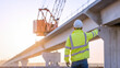 © Hound - A construction worker points to a bridge in the distance. The scene is bright and sunny, with the worker wearing a yellow jacket and a hard hat. The crane in the background is large and orange