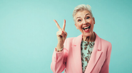 Happy mature woman wearing a pink blazer isolated on a pastel blue background, studio shot of a old retired female celebrating success and win doing a victory V sign gesture with two fingers her hand