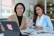 © crizzystudio - Two Asian businesswomen use laptops and smartphones in an open space office. Business concept. Data analysis, roadmap, marketing, accounting, auditing.