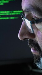 A software developer writing code in close-up. Hacker, cyber security, penetration testing concept. Screen light and computer code reflecting on his face. System administrator. Vertical Format.