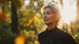 Dojrzała kobieta z uśmiechem na twarzy biegnie przez dziką przyrodę, a słońce tworzy magiczne efekty świetlne, które przenikają przez liście.
