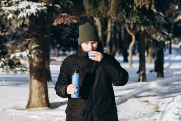 A young bearded man with a thermos enjoys a hot drink in the winter forest. A man drinks tea while walking in the park. Tea from a thermos in the winter forest.