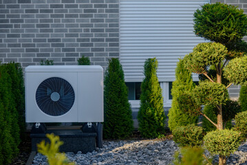 Outdoor heat pump unit set within well-kept garden of trimmed shrubs and stones, placed against brick wall with closed window shutter, forming energy-efficient residential setup. Modern heat pump. 