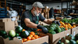 © altitudevisual - Man wearing green apron and cap organizing fresh vegetables and fruits in cardboard boxes at a warehouse setting, focusing on quality produce arrangement.