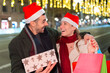 © Raquel - Happy couple in Santa hats sharing gifts and laughter on a decorated street during the holiday season, celebrating and enjoying festive moments.