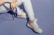 © okrasiuk - Close up female legs in white jeans and retro style high-top multicolor sport sneakers shoes holding sunglasses sitting on purple violet background. Vintage retro fashion style of 80s - 90s vibes