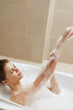 © SHOTPRIME STUDIO - Relaxed young woman enjoying a bubble bath, smiling peacefully while applying foam to her arms. Self-care and wellness concept in a cozy bathroom setting.