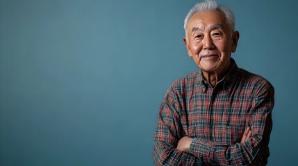 A full body portrait shot of a content and relaxed senior Korean man posed standing against a plain blue studio background dressed in a comfortable plaid button down shirt and slacks