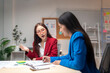 © Wasana - Two women are sitting at a desk, one of them is wearing a red jacket