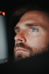 A focused man gazes thoughtfully, illuminated by screens glow, reflecting concentration and determination. His intense blue eyes reveal deep engagement with his work