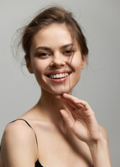 Wall Mural - Cheerful young woman with long hair smiling brightly, hand touching her chin, captured against a neutral grey background. Joy and beauty concept.