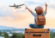 © Quality One   - Child in pilot helmet and goggles points at airplane in sky, sitting on suitcase. scene captures sense of adventure and dreams of flying