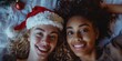 © vefimov - Two smiling girls in Santa hats lying on a festive bedspread during the holiday season.