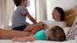 © Marco - Baby lying on a bed with a young boy and mother in the background, enjoying a playful family moment in a relaxed home environment