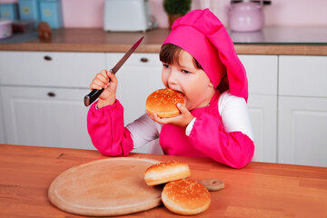 Happy little beautiful girl in a chef costume eats buns while sitting at the kitchen table. Healthy eating.	