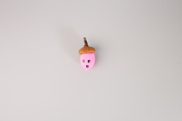  small acorn with a pink painted body and a natural brown cap. decorated to resemble a face, with two black dots for eyes and a small black circle for a mouth. The background is a solid light grey