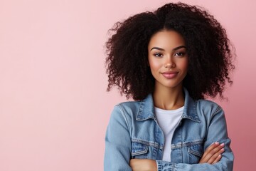 A woman with a curly hairstyle poses in front of a pink background, wearing a denim jacket. Her bright smile and crossed arms exude warmth and approachability.