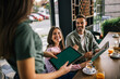 © Mediteraneo - Group of people ordering food from the waitress. Waitress writing orders down on tablet. Undecided people reading the menu while the waitress waits and helps them.