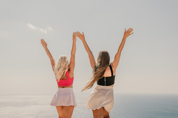 Two women are standing on a beach, one wearing a pink top and the other wearing a black top. They are both smiling and raising their arms in the air, as if they are celebrating something.