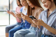 © amnaj - Four young women sitting in a row, engrossed in smartphones, browsing, chatting, and scrolling. Asian friends casually holding devices, enjoying the digital lifestyle
