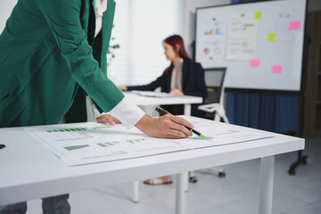 Businesswoman is analyzing financial charts and graphs at a desk in a modern office with a colleague working in the background