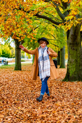 Beautiful brown-haired young woman wearing beige coat, jeans, checkered warm scarf, black brimmed hat walking along alley with old trees in city park on autumn day. Front view