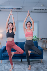 Two young asian women are practicing yoga together in their living room, maintaining balance and focus in the tree pose, promoting wellness and mindfulness