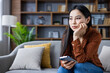 © Liubomir - Young Asian woman sits on couch, holding phone, looking thoughtful. Casual attire suggests relaxed setting. Expression indicates contemplation or sadness, perhaps personal reflection at home.