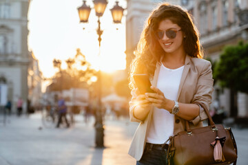 Wall Mural - A fashionable mid-adult Caucasian woman, in chic streetwear, expertly navigates her smartphone against a bustling city backdrop, bathed in the warm glow of sunset.