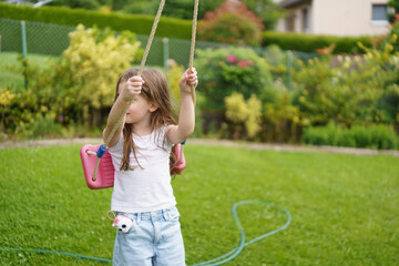 A little cheerful girl without one front tooth dressed in a T-shirt and jeans is riding on a swing in the backyard of her house. 