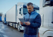 © iamfrk7 - Middle-aged man using a tablet outdoors near trucks, concept of modern logistics and transport management, checking data in transport industry