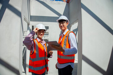 Two construction workers wearing orange vests and hard hats are standing in front of a building. One of them is holding a tablet