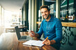 © peopleimages.com - Remote work, happy man and portrait in cafe with phone, writing and multitask for startup management at laptop. Entrepreneur, smile and tech at table for planning with calendar update in restaurant