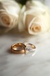 © Tcukimay - Two gold and white wedding bands with diamonds sit on a white marble surface with two white roses blurred in the background.