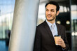 © Minerva Studio - Confident young businessman in a suit enjoys a coffee break outside a modern office building, exemplifying corporate lifestyle and successful professional routine