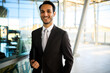 © Minerva Studio - Confident male executive smiling in a tailored suit, holding a tablet, standing in a modern office with a glass backdrop