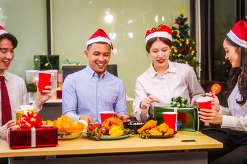 A festive office Christmas party with friends and colleagues celebrating after work. Asian business people wear Santa hats, enjoy grilled chicken, drinks, and gifts, indoor with holiday lights.