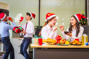 A festive office Christmas party with friends and colleagues celebrating after work. Asian business people wear Santa hats, enjoy grilled chicken, drinks, and gifts, indoor with holiday lights.