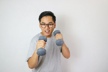 Asian man holds two gray dumbbells in a boxing style, wearing a gray t-shirt. He stands against a white background, showing focus during his strength training.